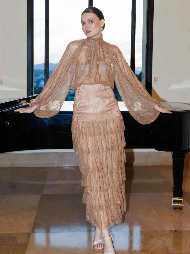 Model in a caramel lace gown with sheer bishop sleeves and a pussy-bow neckline, hands resting on a black grand piano