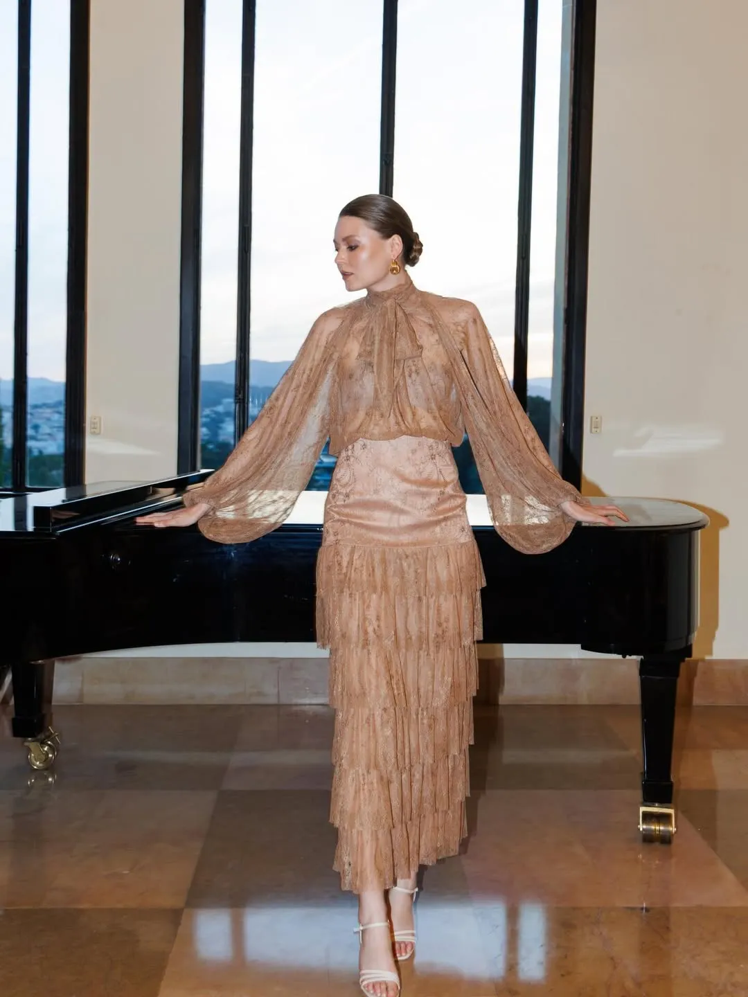 Full-length caramel lace gown with tiered skirt, arms resting on the grand piano