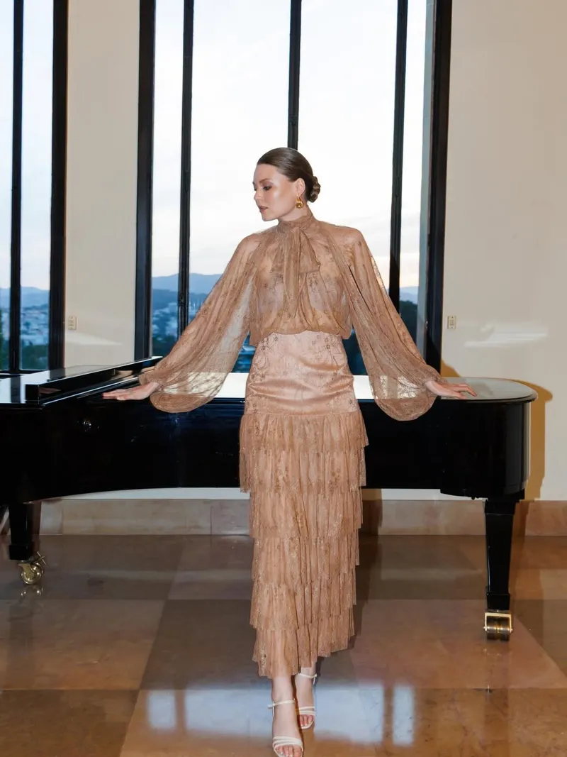 Full-length caramel lace gown with tiered skirt, arms resting on the grand piano