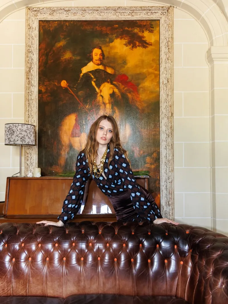 Model leaning on the back of a brown leather chesterfield beneath a large equestrian oil painting, wearing the diamond-print wrap blouse and gold chain necklaces