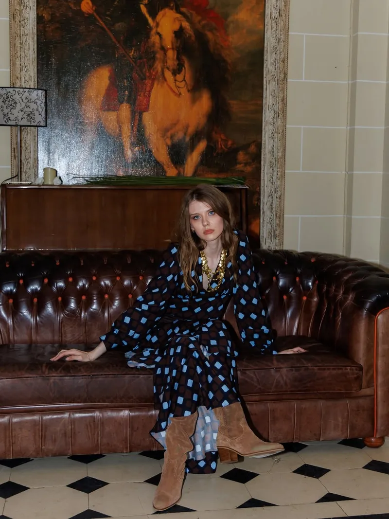 Model seated on a chesterfield sofa on a black and white tiled floor in the diamond-print wrap dress look and tan suede boots