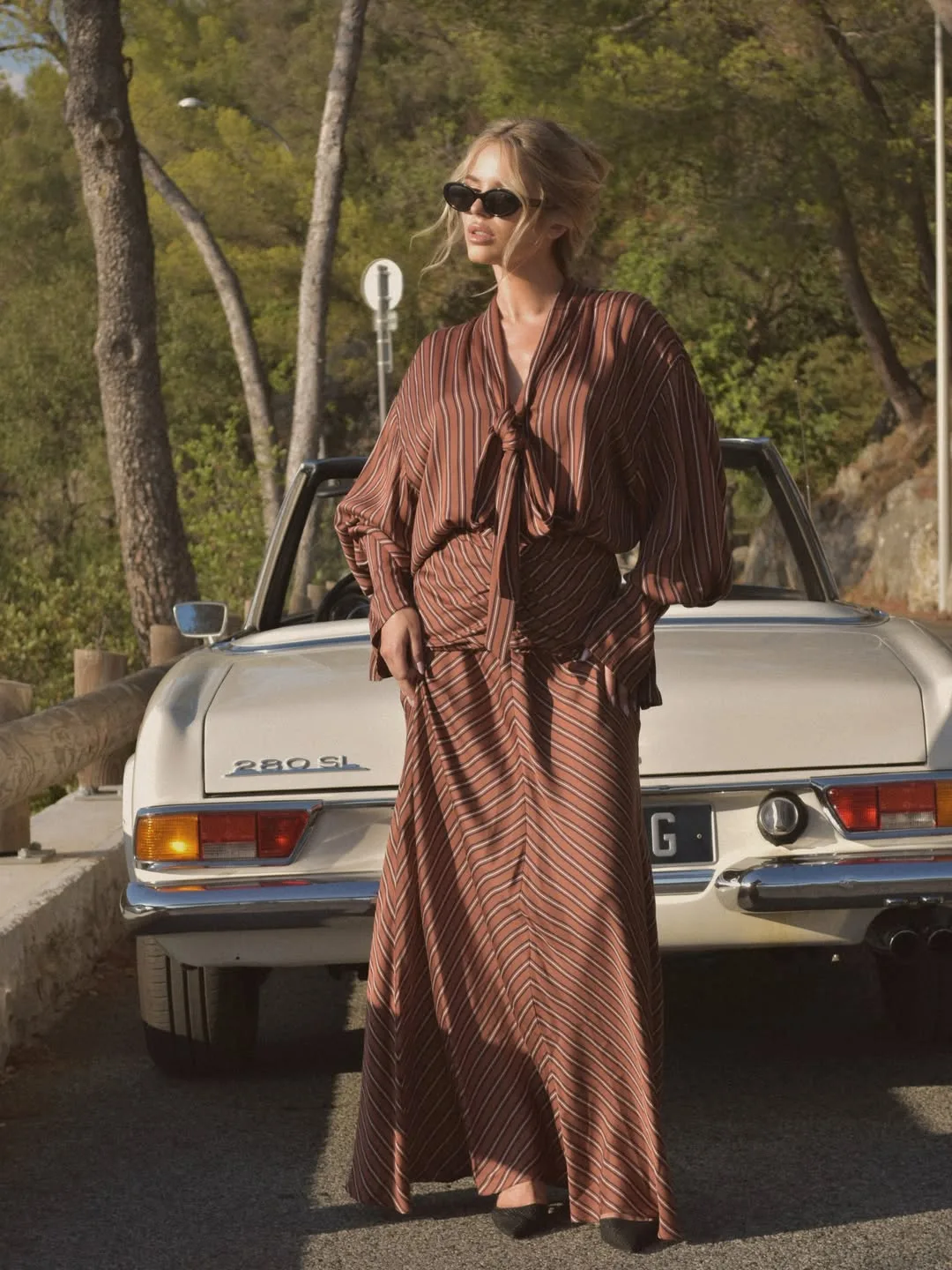 Model standing in front of a cream vintage convertible on a pine-lined coast road, in a rust striped tie-neck blouse and matching bias-cut maxi skirt
