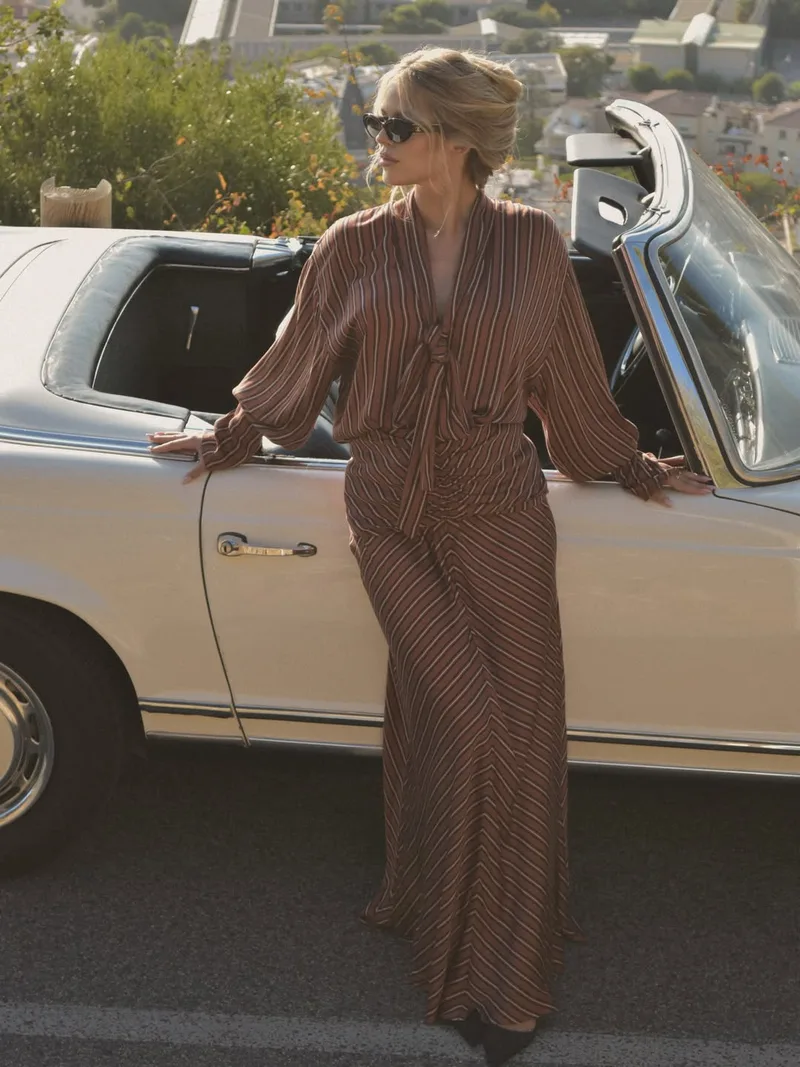 Model leaning against the side of the convertible in the striped set, the town and hills behind her