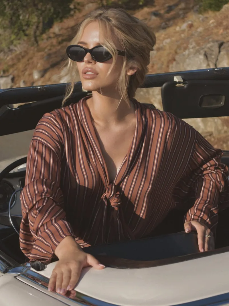 Close portrait in the open car: the knotted tie-neck of the rust striped blouse and oval black sunglasses