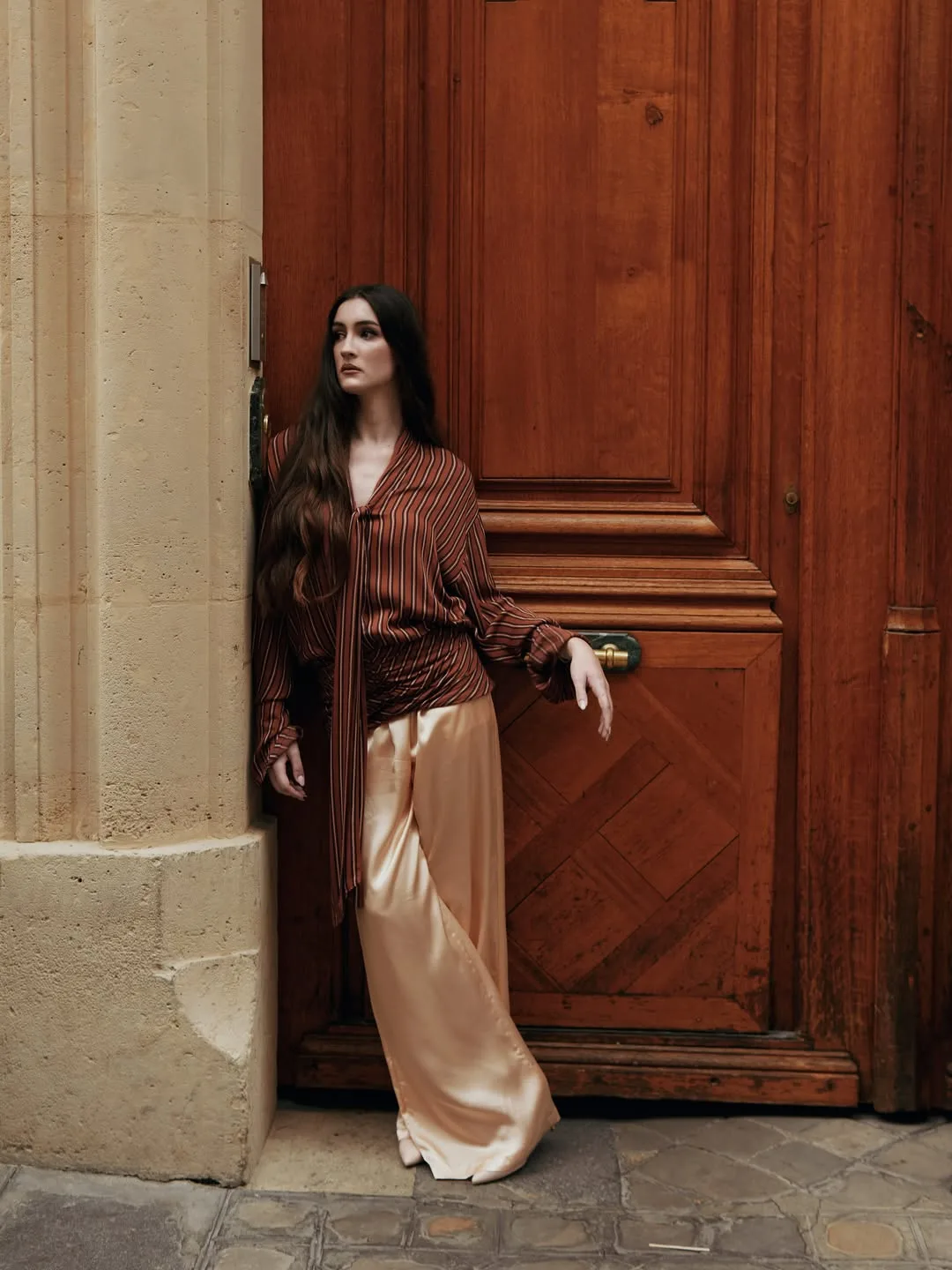 Model leaning in a stone doorway against a carved oak door in a rust striped tie-neck blouse and wide-leg champagne satin trousers