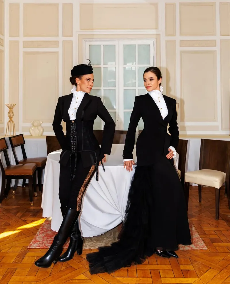 Two models in black equestrian tailoring with white ruffled high-neck blouses in a panelled salon
