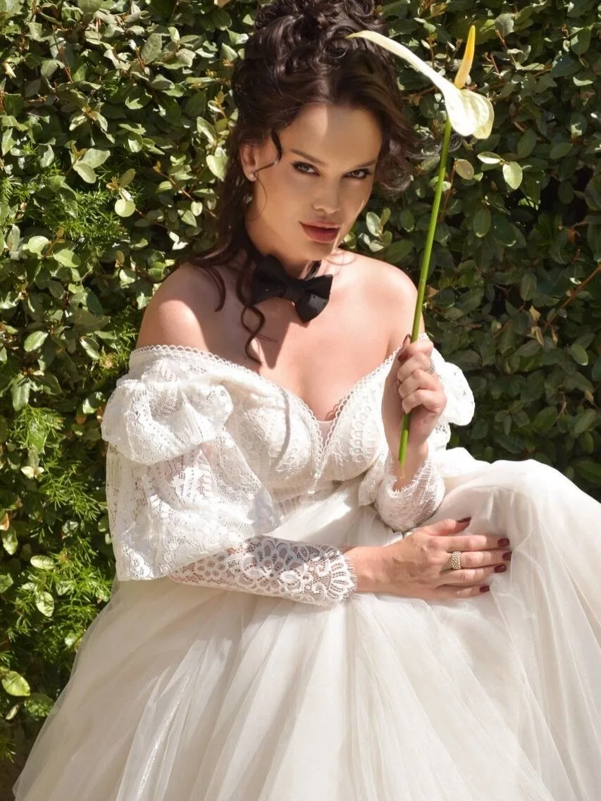 Model in an ivory lace off-shoulder gown with a black bow tie, holding a white calla lily against a green hedge