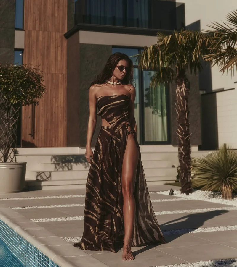 Model walking beside a pool in a strapless striped bandeau top and a high-split wrap maxi skirt, wearing a shell necklace and sunglasses